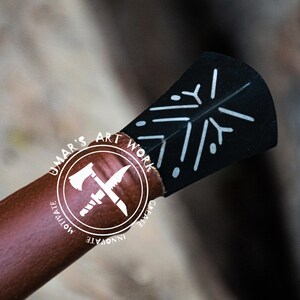 May include: Close-up of a handcrafted axe head with a black blade featuring a white snowflake design. The axe handle is wrapped in brown leather. The image includes the text "UMAR'S ART WORK" in a circular logo.