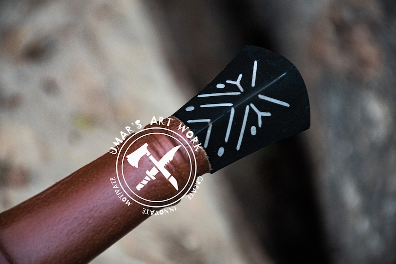 May include: Close-up of a handcrafted axe head with a black blade featuring a white snowflake design. The axe handle is wrapped in brown leather. The image includes the text "UMAR'S ART WORK" in a circular logo.
