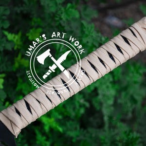 K&ouml;nnte beinhalten: Nahaufnahme einer schwarzen Klinge mit einem beige umwickelten Griff. Der Griff ist in einem Kreuzmuster gewickelt. Der Text "UMAR'S ART WORK" befindet sich in einem wei&szlig;en Kreis mit einem gekreuzten Axt- und Messerdesign. Der Hintergrund ist verschwommenes gr&uuml;nes Laub.