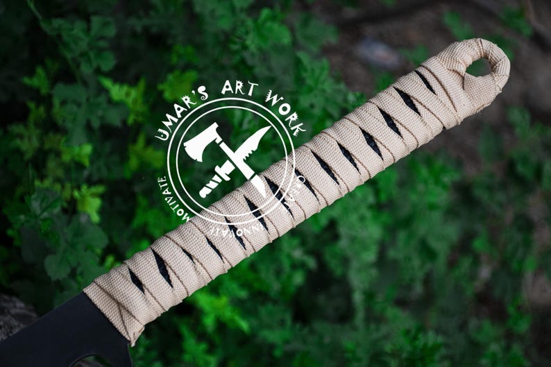 K&ouml;nnte beinhalten: Nahaufnahme einer schwarzen Klinge mit einem beige umwickelten Griff. Der Griff ist in einem Kreuzmuster gewickelt. Der Text "UMAR'S ART WORK" befindet sich in einem wei&szlig;en Kreis mit einem gekreuzten Axt- und Messerdesign. Der Hintergrund ist verschwommenes gr&uuml;nes Laub.