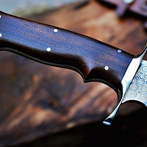 Puede incluir: Primer plano de un cuchillo artesanal con mango de madera oscura y hoja de acero estampada. El mango presenta incrustaciones de puntos blancos. La hoja tiene un patr&oacute;n arremolinado &uacute;nico. Cuchillo para actividades al aire libre.