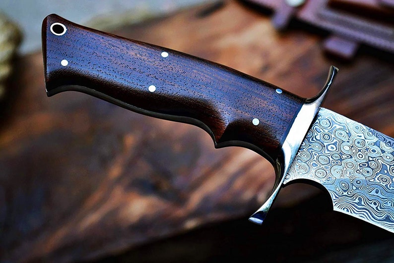 Puede incluir: Primer plano de un cuchillo artesanal con mango de madera oscura y hoja de acero estampada. El mango presenta incrustaciones de puntos blancos. La hoja tiene un patr&oacute;n arremolinado &uacute;nico. Cuchillo para actividades al aire libre.