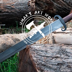 K&ouml;nnte beinhalten: Ein schwarzes Schwert mit einem Holzgriff und einem dekorativen Metallknauf. Das Schwert liegt auf einem Baumstamm mit einem wei&szlig;en Kreislogo, auf dem "Umar's Art Work" steht, und den Worten "Innovate" und "Motivate" auf beiden Seiten des Logos.