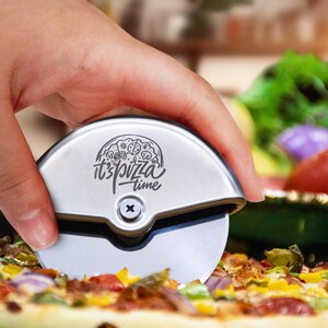 May include: A silver-coloured circular pizza cutter with a metallic handle featuring a black engraved graphic of a pizza and the text "it's pizza time" above a central screw. The tool has a sharp, rotating circular blade for slicing pizza.