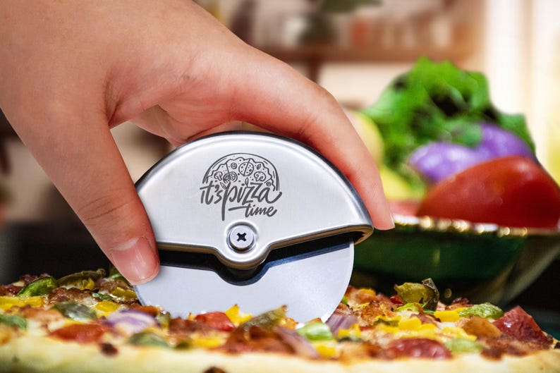 May include: A silver-coloured circular pizza cutter with a metallic handle featuring a black engraved graphic of a pizza and the text "it's pizza time" above a central screw. The tool has a sharp, rotating circular blade for slicing pizza.