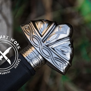 May include: Close-up of a sword pommel with intricate silver and black Celtic knotwork designs. The pommel is made of metal and has a polished finish. The sword is part of a decorative display.