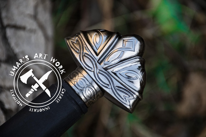 May include: Close-up of a sword pommel with intricate silver and black Celtic knotwork designs. The pommel is made of metal and has a polished finish. The sword is part of a decorative display.