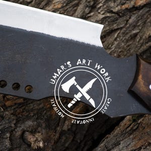 K&ouml;nnte beinhalten: Ein gro&szlig;es, schwarzes Metallbeil mit einer silbernen Klinge und einem dunklen Holzgriff. Das Beil hat eine einzigartige Form und tr&auml;gt den Text "UMAR'S ART WORK" in einem kreisf&ouml;rmigen Logo. Die Klinge hat drei L&ouml;cher in der N&auml;he des Griffs.