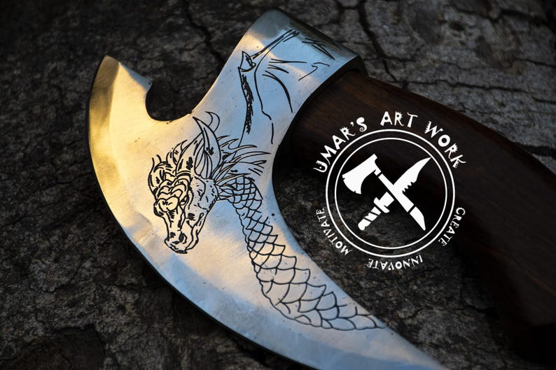 May include: A metal axe head with a dark wooden handle featuring an etched dragon design on the blade. A circular logo is visible on the blade with the text UMAR'S ART WORK, MOTIVATE, CREATE, INNOVATE, and an icon of an axe and a knife crossed.