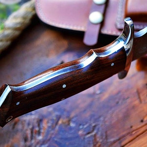 Puede incluir: Primer plano de un cuchillo de caza con mango de madera oscura y refuerzo plateado. La hoja es parcialmente visible y el cuchillo descansa sobre una superficie de madera. Una funda de cuero est&aacute; en el fondo.