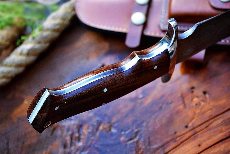 Puede incluir: Primer plano de un cuchillo de caza con mango de madera oscura y refuerzo plateado. La hoja es parcialmente visible y el cuchillo descansa sobre una superficie de madera. Una funda de cuero est&aacute; en el fondo.