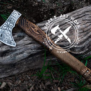 Op de afbeelding: Een handgemaakte bijl met een houten steel en een stalen kop. De kop is versierd met ingewikkelde houtsnijwerk, waaronder een Viking-knoopwerkontwerp. De steel is omwikkeld met bruin leer en heeft een metalen dop aan het einde. De bijl rust op een stuk hout met de tekst "UMAR'S ART WORK" in een cirkel rond twee gekruiste bijlen.