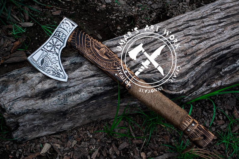 Op de afbeelding: Een handgemaakte bijl met een houten steel en een stalen kop. De kop is versierd met ingewikkelde houtsnijwerk, waaronder een Viking-knoopwerkontwerp. De steel is omwikkeld met bruin leer en heeft een metalen dop aan het einde. De bijl rust op een stuk hout met de tekst "UMAR'S ART WORK" in een cirkel rond twee gekruiste bijlen.