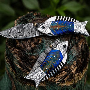 May include: Two fish-shaped pocket knives with Damascus steel blades and handles. The handles feature a blue and brown design with white and black accents. The knives are displayed on a weathered wooden surface against a blurred green background.