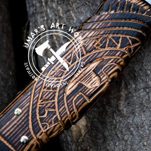 May include: Close-up of a wooden axe handle with intricate carvings. The handle is dark brown with black accents and features a repeating pattern of geometric shapes. The text "UMAR'S ART WORK" is visible on the handle.