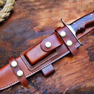 Puede incluir: Un cuchillo de caza artesanal con mango de madera oscura y hoja de acero inoxidable, asegurado en una funda de cuero marr&oacute;n. La funda tiene un lazo para cintur&oacute;n y se sujeta con remaches color lat&oacute;n. El cuchillo descansa sobre una superficie de madera.