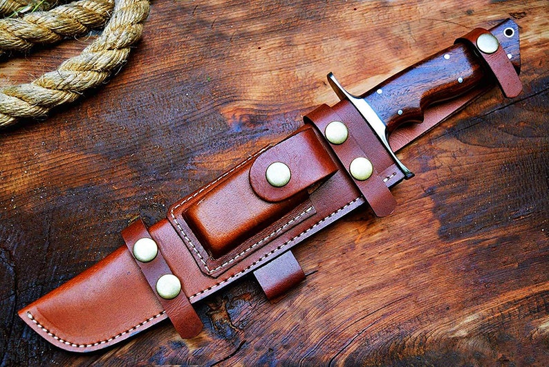 Puede incluir: Un cuchillo de caza artesanal con mango de madera oscura y hoja de acero inoxidable, asegurado en una funda de cuero marr&oacute;n. La funda tiene un lazo para cintur&oacute;n y se sujeta con remaches color lat&oacute;n. El cuchillo descansa sobre una superficie de madera.