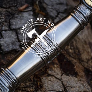 May include: Close-up of a silver sword hilt with intricate details and a gold-colored accent. The hilt is decorated with a repeating pattern of stylized leaves and flowers. The sword is resting on a rough wooden surface. The text "UMAR'S ART WORK" is visible on the hilt.