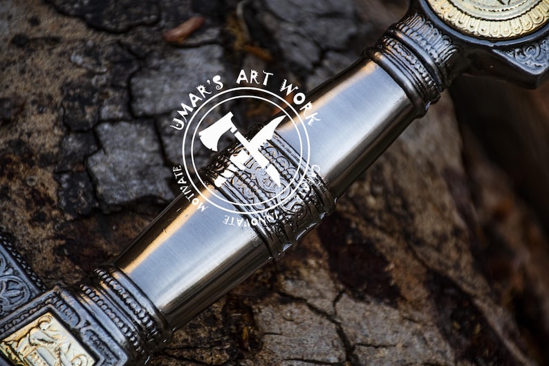 May include: Close-up of a silver sword hilt with intricate details and a gold-colored accent. The hilt is decorated with a repeating pattern of stylized leaves and flowers. The sword is resting on a rough wooden surface. The text "UMAR'S ART WORK" is visible on the hilt.