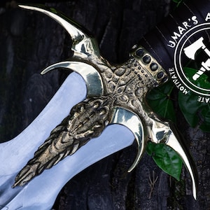 Könnte beinhalten: Nahaufnahme eines Fantasy-Dolches mit silberner Klinge und aufwändigem goldfarbenem Griff. Der Griff weist komplizierte Designs und gebogene Elemente auf. Das Bild enthält den Text "UMAR'S ART WORK" in einem kreisförmigen Logo.