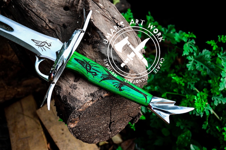 May include: A silver sword with a green and black wooden handle. The handle has a decorative design and the text "UMAR'S ART WORK CREATE INNOVATE MOTIVATE" is on the handle.