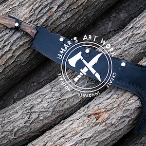 K&ouml;nnte beinhalten: Eine handgefertigte Axt mit Holzgriff und schwarzer Klingenh&uuml;lle. Die Axt hat einen dunklen Holzgriff und eine schwarze Klingenh&uuml;lle mit dem Text "UMAR'S ART WORK". Die Axt liegt auf einem Stapel Holzst&auml;mme.