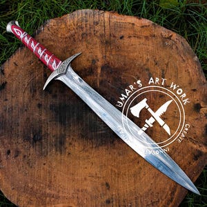 May include: A handcrafted sword with a silver blade and a red wrapped handle, resting on a wooden surface. The sword features intricate designs and is displayed on a wooden slab with the text "UMAR'S ART WORK".