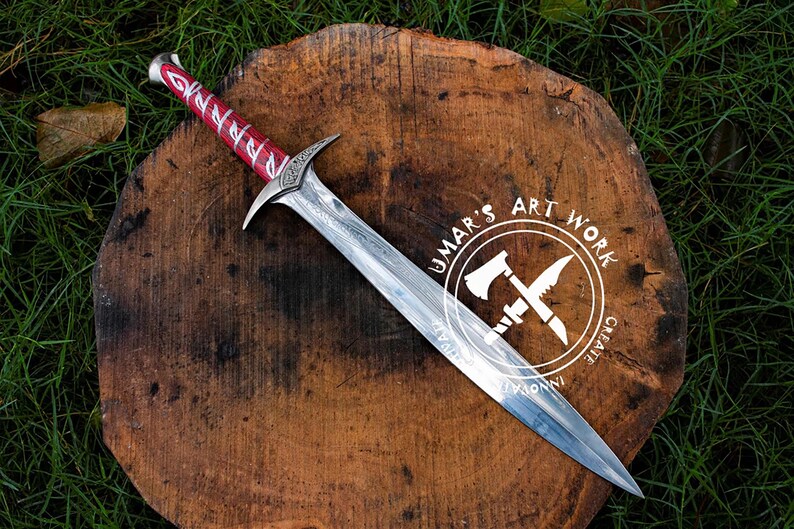 May include: A handcrafted sword with a silver blade and a red wrapped handle, resting on a wooden surface. The sword features intricate designs and is displayed on a wooden slab with the text "UMAR'S ART WORK".