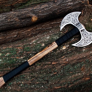 Könnte beinhalten: Eine doppelseitige Axt mit einem Holzstiel und einer schwarzen Lederummantelung. Die Axtköpfe sind mit komplizierten keltischen Knotenmustern verziert.