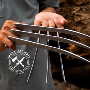 Può includere: Artigli in metallo argentato con un design a lama curva, tenuti davanti a una persona. Gli artigli sono il punto focale, con un logo che dice "Umar's Art Work" visibile sulla camicia della persona.