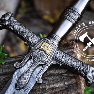 May include: A detailed, intricately carved sword with a silver blade and gold accents. The hilt features a cross guard with ornate designs and a decorative pommel. The sword is resting on a wooden surface. The image includes the text "UMAR'S ART WORK CREATE INNOVATE MOTIVATE".