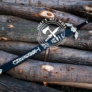 May include: A black sword with silver accents and a decorative design, resting on a pile of logs. The sword features a detailed hilt and blade design. The image includes a circular logo with text that reads "UMAR'S ART WORK".