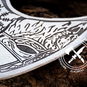 May include: A close-up of a silver axe head with a black and white design. The design features a stylized tree with a bird and a geometric pattern. The axe head is resting on a piece of wood.