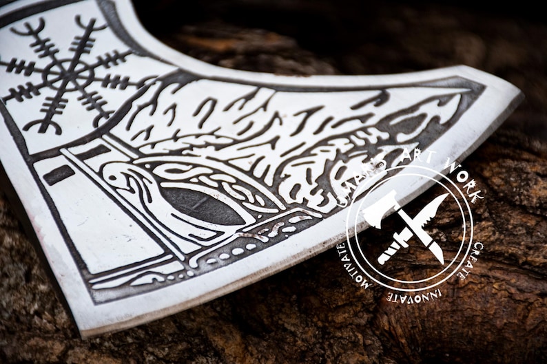 May include: A close-up of a silver axe head with a black and white design. The design features a stylized tree with a bird and a geometric pattern. The axe head is resting on a piece of wood.
