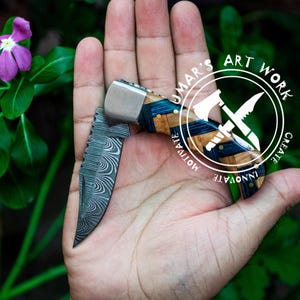 May include: A pocket knife with a blue and wood handle. The blade is made of Damascus steel and has a wavy pattern. The knife is open and held in a hand. The knife has the words "UMAR'S ART WORK" and "CREATE INNOVATE MOTIVATE" on the handle.