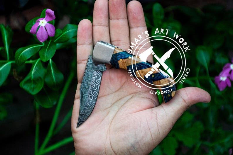 May include: A pocket knife with a blue and wood handle. The blade is made of Damascus steel and has a wavy pattern. The knife is open and held in a hand. The knife has the words "UMAR'S ART WORK" and "CREATE INNOVATE MOTIVATE" on the handle.