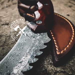 Puede incluir: Primer plano de un cuchillo personalizado con una hoja estampada y un mango de madera. El cuchillo est&aacute; parcialmente enfundado en una funda de cuero marr&oacute;n con costuras visibles. La hoja tiene un patr&oacute;n ondulado &uacute;nico. El texto "UMAR'S ART WORK" es visible.