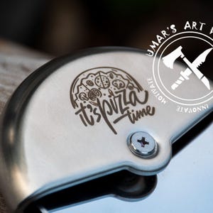 May include: A stainless steel pizza cutter featuring a circular blade and a protective handle. The handle is engraved with a graphic of a pizza slice and the text "it's pizza time". A circular logo with an axe and knife icon reads "UMAR'S ART WORK".