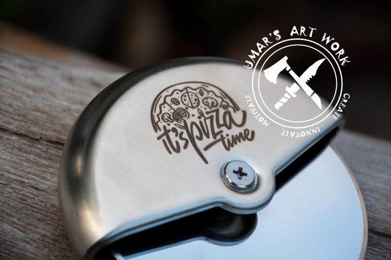 May include: A stainless steel pizza cutter featuring a circular blade and a protective handle. The handle is engraved with a graphic of a pizza slice and the text "it's pizza time". A circular logo with an axe and knife icon reads "UMAR'S ART WORK".