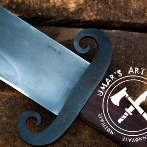 K&ouml;nnte beinhalten: Nahaufnahme eines handgefertigten Messers mit schwarzem Griff und silberner Klinge. Der Griff hat ein einzigartiges Design mit zwei Schlaufen. Das Messer liegt auf einer Holzoberfl&auml;che. Das Bild enth&auml;lt auch ein Logo mit dem Text "UMAR'S ART WORK" und den W&ouml;rtern "INNOVATE", "CREATE" und "MOTIVATE" um das Logo herum.