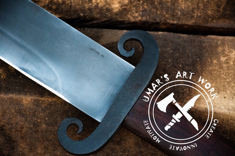 K&ouml;nnte beinhalten: Nahaufnahme eines handgefertigten Messers mit schwarzem Griff und silberner Klinge. Der Griff hat ein einzigartiges Design mit zwei Schlaufen. Das Messer liegt auf einer Holzoberfl&auml;che. Das Bild enth&auml;lt auch ein Logo mit dem Text "UMAR'S ART WORK" und den W&ouml;rtern "INNOVATE", "CREATE" und "MOTIVATE" um das Logo herum.