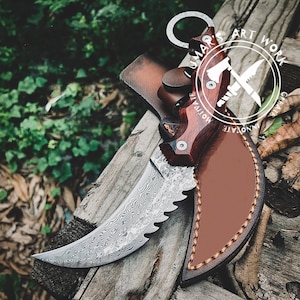 Puede incluir: Un cuchillo de acero de Damasco con una hoja curva y un borde dentado, con un mango de madera oscura y una funda de cuero. El cuchillo tiene un anillo al final del mango. La imagen incluye el texto "LUMAR'S ART WORK".