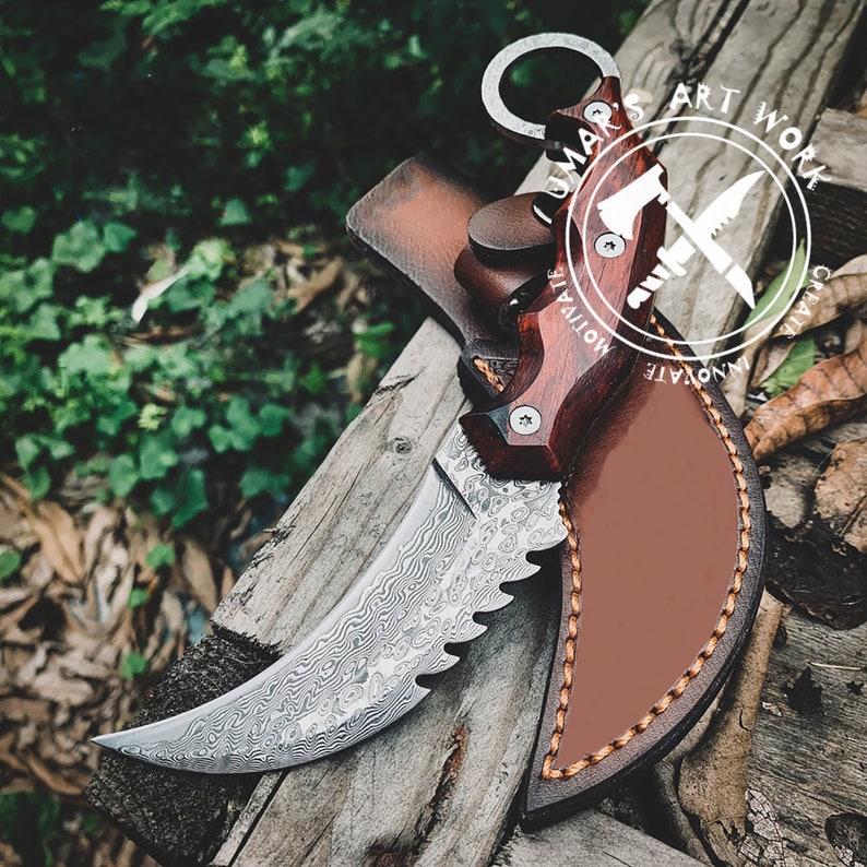Puede incluir: Un cuchillo de acero de Damasco con una hoja curva y un borde dentado, con un mango de madera oscura y una funda de cuero. El cuchillo tiene un anillo al final del mango. La imagen incluye el texto "LUMAR'S ART WORK".