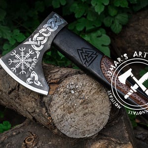 Op de afbeelding: Een zwarte en zilveren bijl met een houten steel. De bijlkop is versierd met een Keltisch knoopontwerp en een Viking-symbool. De steel is versierd met een Viking-symbool en de woorden "UMAR'S ART WORK CREATE INNOVATE MOTIVATE".