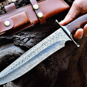 Puede incluir: Un cuchillo de acero de Damasco con una hoja estampada y un mango de madera oscura. El cuchillo tiene una virola plateada y va acompa&ntilde;ado de una funda de cuero marr&oacute;n con cierres de bot&oacute;n a presi&oacute;n. El cuchillo se sostiene en una mano.