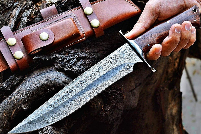 Puede incluir: Un cuchillo de acero de Damasco con una hoja estampada y un mango de madera oscura. El cuchillo tiene una virola plateada y va acompa&ntilde;ado de una funda de cuero marr&oacute;n con cierres de bot&oacute;n a presi&oacute;n. El cuchillo se sostiene en una mano.