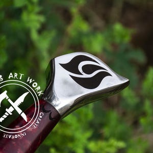 May include: Close-up of a silver sword pommel with a black flame design. The pommel is attached to a red and black wooden handle with the text "UMAR'S ART WORK CREATE INNOVATE MOTIVATE" in white.