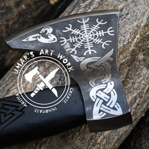 Op de afbeelding: Een zwarte bijlkop met een zilveren steel. De bijlkop is versierd met verschillende Noorse symbolen, waaronder een Vegvisir, een knoopwerkontwerp en een triskelion. De steel is gegraveerd met de woorden "Umar's Art Work, Create, Innovate, Motivate".