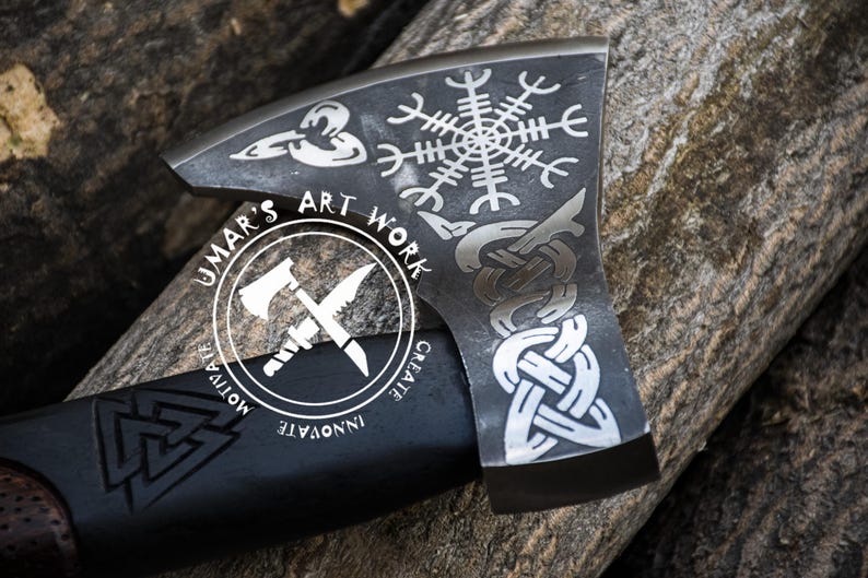 Op de afbeelding: Een zwarte bijlkop met een zilveren steel. De bijlkop is versierd met verschillende Noorse symbolen, waaronder een Vegvisir, een knoopwerkontwerp en een triskelion. De steel is gegraveerd met de woorden "Umar's Art Work, Create, Innovate, Motivate".