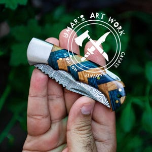 May include: A pocket knife with a blue and brown wood handle and a stainless steel blade. The blade has a wavy pattern and a serrated edge. The knife is closed and being held in a hand. The knife has the words "UMAR'S ART WORK CREATE INNOVATE" on the handle.